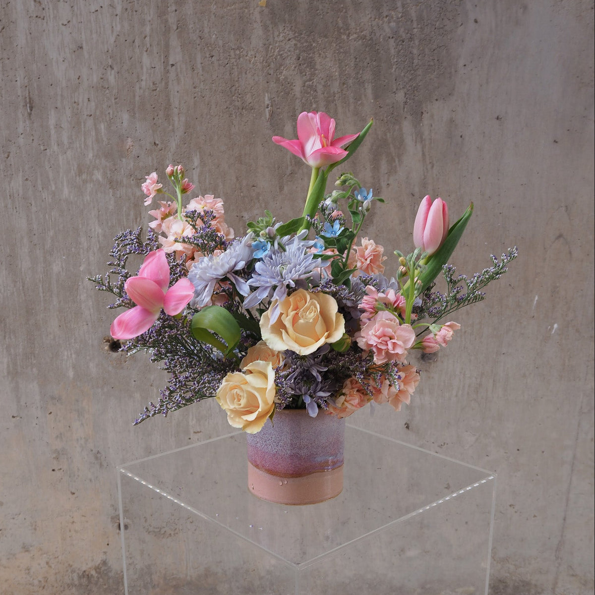 A pastel floral arrangement in a small pink and purple vase, displayed on a clear acrylic pedestal against a textured concrete wall. The bouquet includes pink tulips, pale yellow roses, peach carnations, lavender-blue hydrangeas, and green foliage.