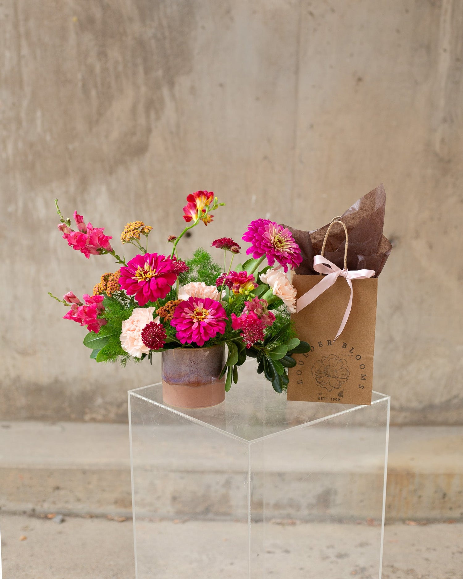 A vibrant flower arrangement in a small mauve pot next to a brown kraft paper gift bag with a pink ribbon, displayed on a clear acrylic pedestal against a textured concrete wall.